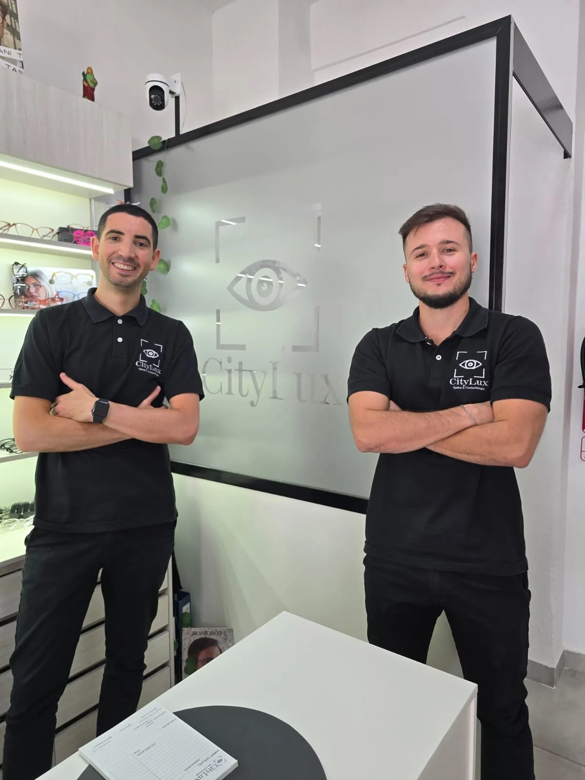 Los dos ópticos de CityLux de pie en el local, de brazos cruzados, delante del logo